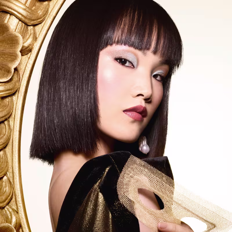 Apresenta uma modelo com maquiagem impactante, incluindo sombras nos olhos em tons dourados e marrons, delineador marcante e batom vermelho escuro. Ela tem cabelos lisos e curtos na altura do queixo, com uma franja reta.