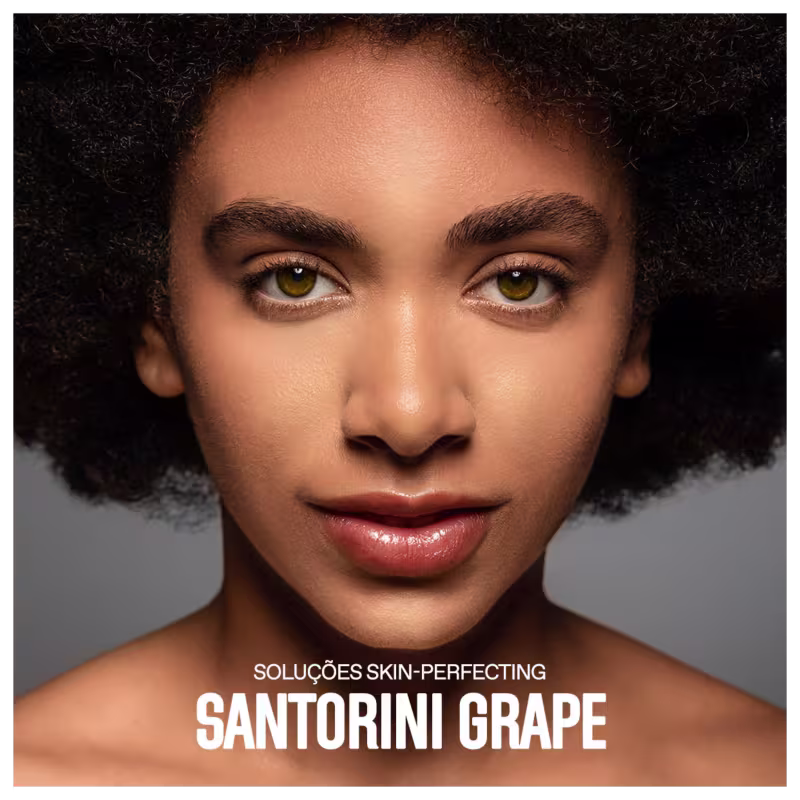 A imagem mostra um close-up do rosto de uma mulher negra com cabelos crespos e curtos. Ela tem olhos amendoados de cor amarelo-esverdeada. Sua pele tem um aspecto aveludado e uniforme, provavelmente resultado do uso dos produtos anunciados.