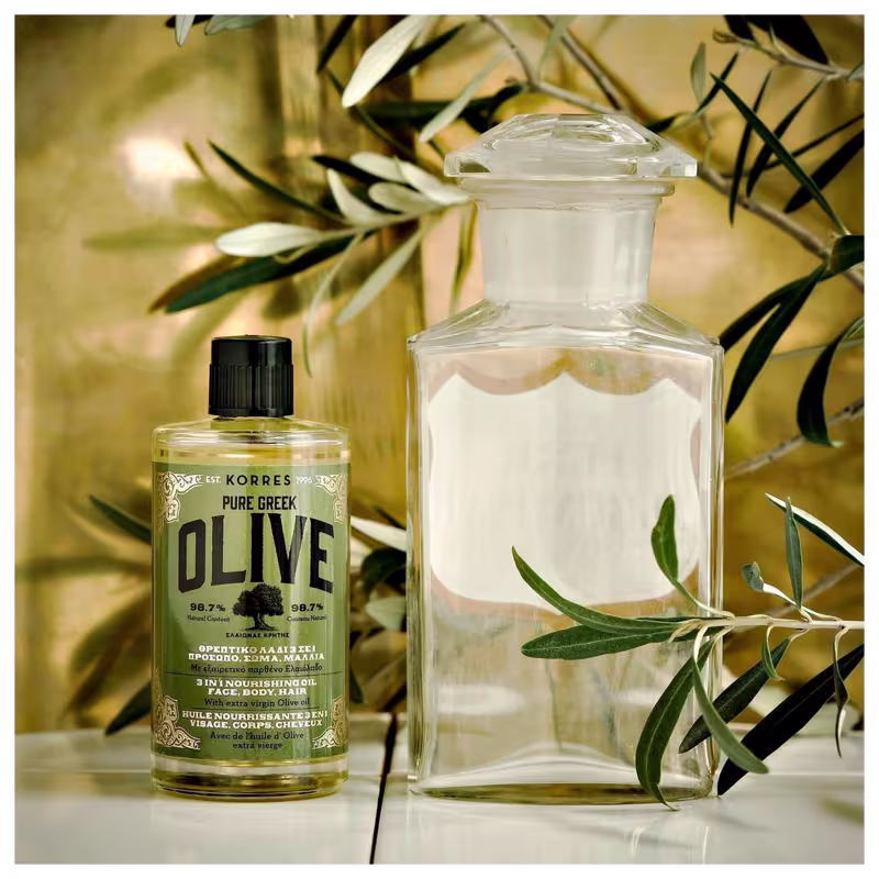 A imagem mostra o produto e ao lado um frasco de vidro transparente. Ambos apresentam galhos de oliveira ao redor, simbolizando os ingredientes naturais. As cores predominantes são transparente, verde oliva e preto.