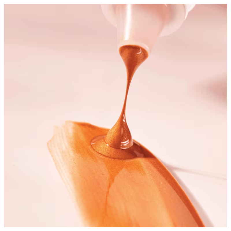 O bronzer está sendo despejado de um frasco ou recipiente, formando um filete ou gota alongada e pontiaguda. Destacando sua textura sedosa e brilhante contra um fundo desfocado em tons claros e suaves.