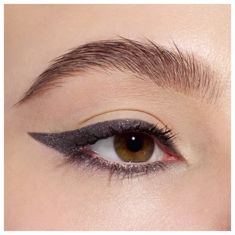 A imagem mostra uma maquiagem de olhos com um delineado gatinho dramático em glitter chumbo cintilante. O delineado é alongado e afiado nas extremidades externas, criando um efeito marcante. A sombra é suave e brilhante, realçando os olhos castanhos.