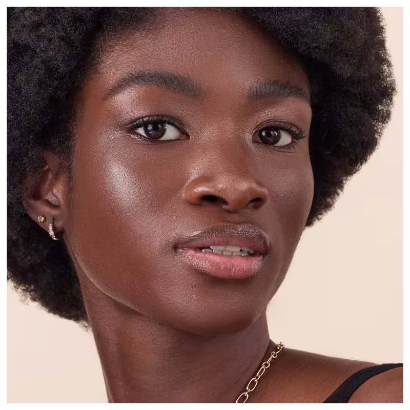 Jovem mulher negra com cabelos crespos e curtos. Ela tem olhos castanhos expressivos, sobrancelhas bem delineadas e lábios cheios levemente brilhantes. Sua pele é aveludada e brilhante, com um tom marrom profundo e saudável.