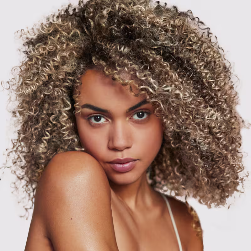 A imagem mostra uma mulher com cabelos cacheados e loiros, destacando o efeito de leveza e brilho do óleo Kérastase.