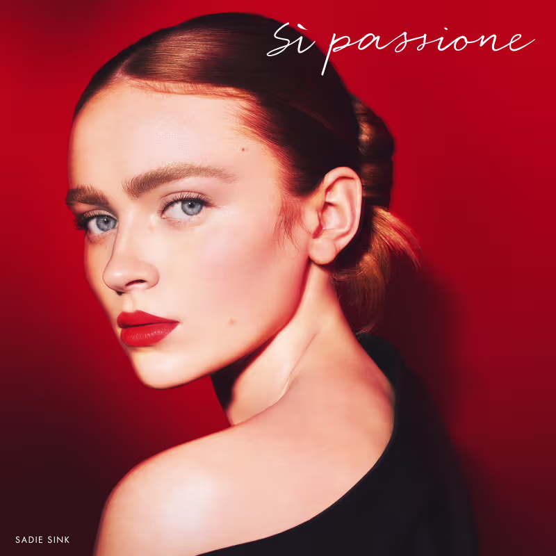A imagem mostra a atriz Sadie Sink bem maquiada e com o cabelo preso. Sua expressão é intensa, os lábios estão com um batom vermelho da mesma cor do fundo da foto.