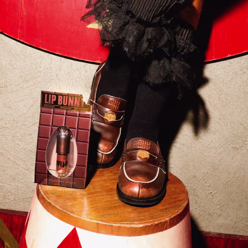 Gloss Labial Fran by Franciny Ehlke Lip Bunny ao lado de sapatos tipo mocassim na cor marrom