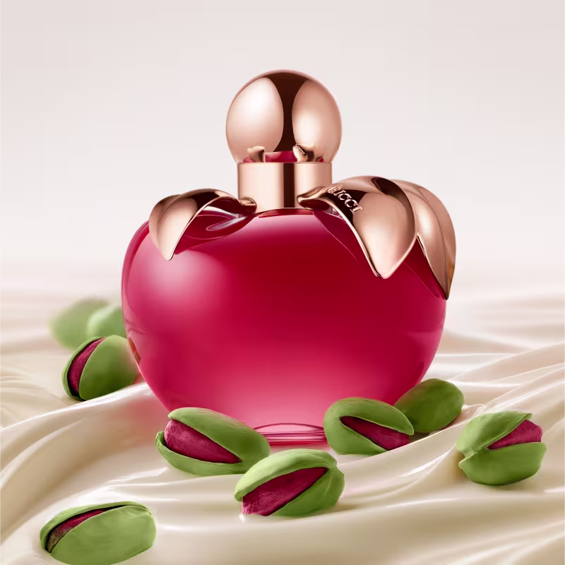 Frasco de perfume vermelho com detalhes dourados em formato de maçã, rodeado por pétalas verdes, em um cenário com tecido branco suave.