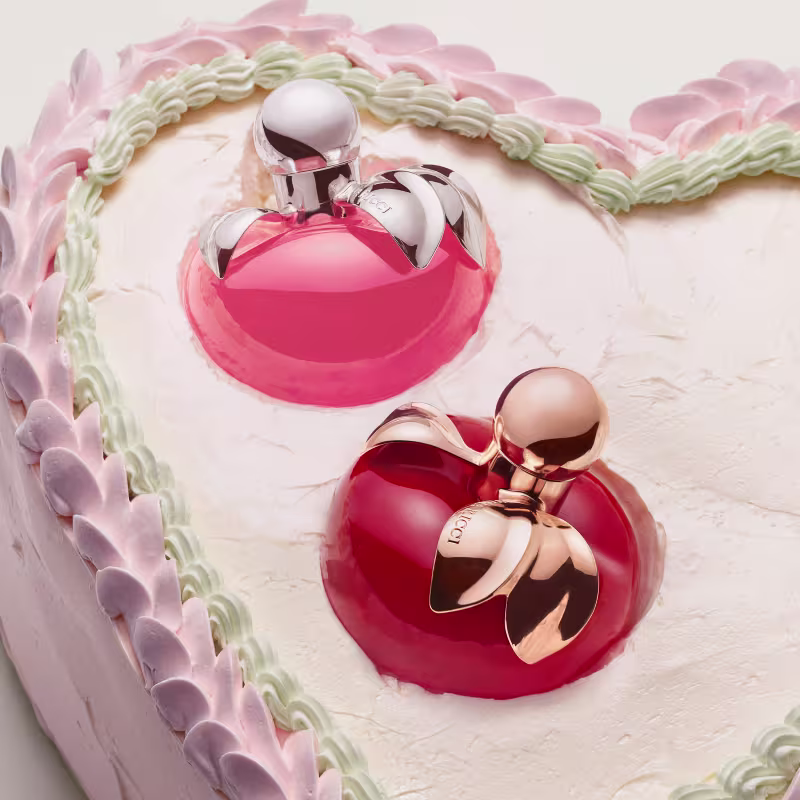 Dois frascos de perfume feminino com embalagens em tons de rosa e vermelho, com detalhes em metal prateado. O frasco maior possui um laço decorativo na tampa. Ambos os produtos exibem um design elegante e sofisticado e estão no topo de um bolo.