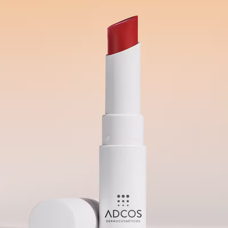 Bálsamo Hidratante Labial com embalagem branca com tampa e logotipo da marca Adcos Dermocosmética em cinza. O bálsamo possui uma cor vermelha escura e brilhante.