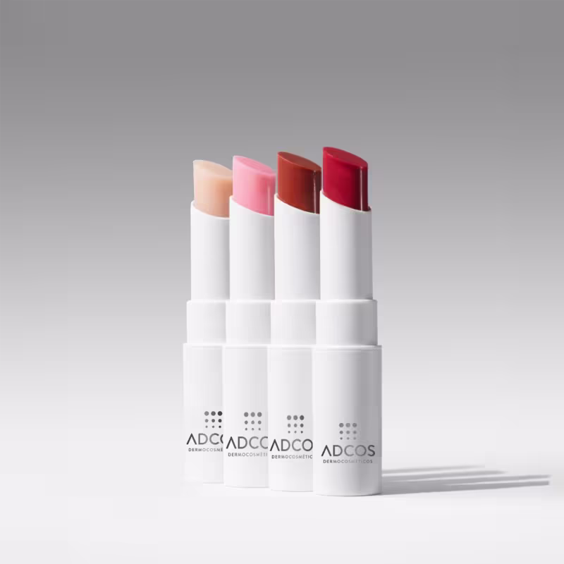 Vários bálsamos em tons de vermelho e rosa, com embalagens brancas e cilíndricas, dispostos em um fundo branco.