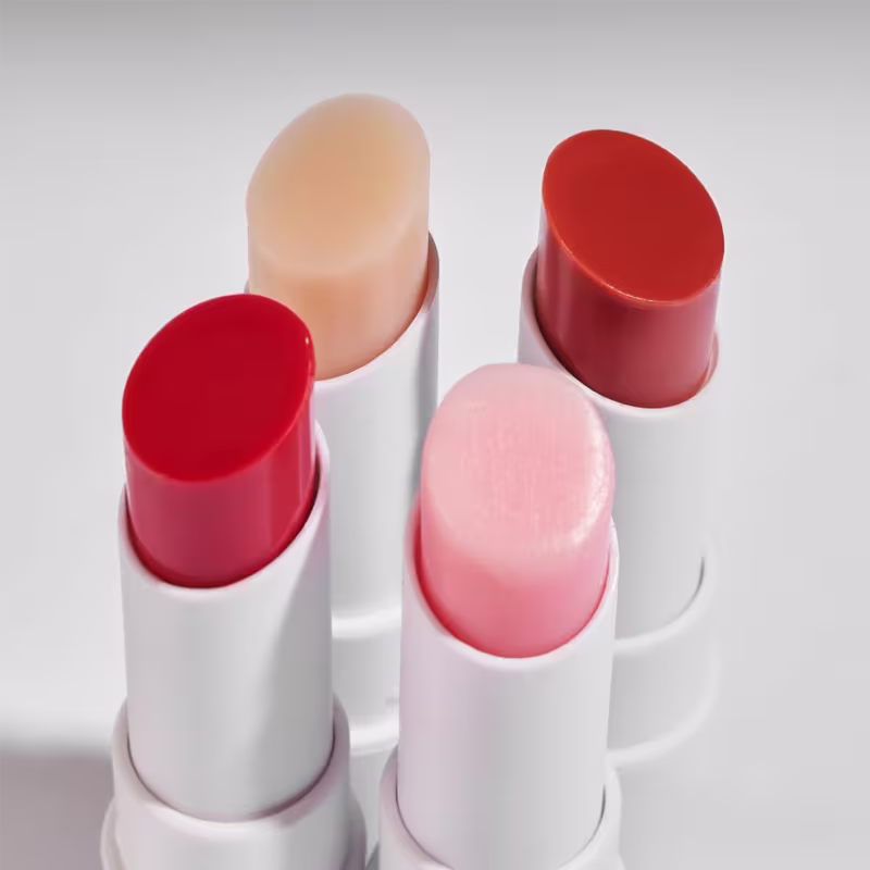 Vários bálsamos em tons de vermelho e rosa, com embalagens brancas e cilíndricas, dispostos em um fundo branco.