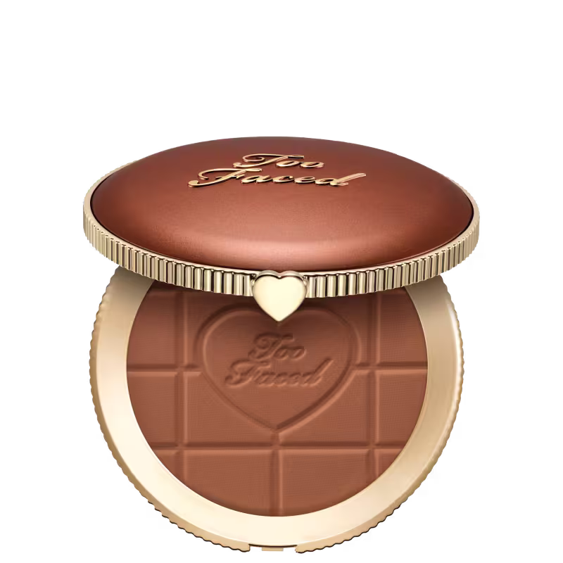 Pó bronzeador compacto com acabamento aveludado em embalagem redonda de metal dourado com detalhes em relevo e coração no centro. Apresenta textura e tonalidade marrom escura com inscrição "Too Faced" na superfície.
