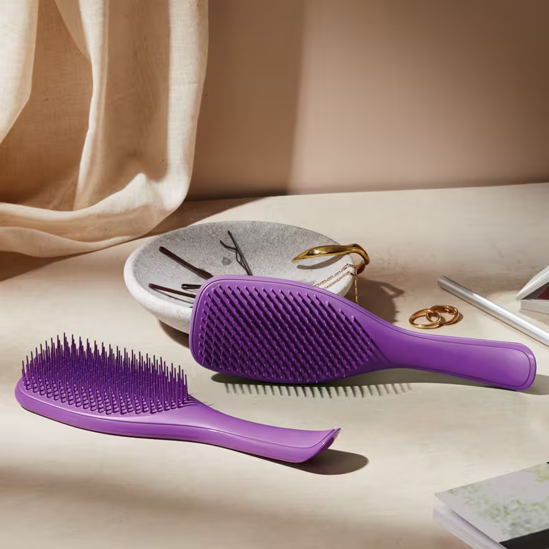 Ambiente de penteado e cuidados pessoais com acessórios em tons neutros, destacando-se a escova de cabelo da marca Tangle Teezer em cor roxa, posicionada em primeiro plano.