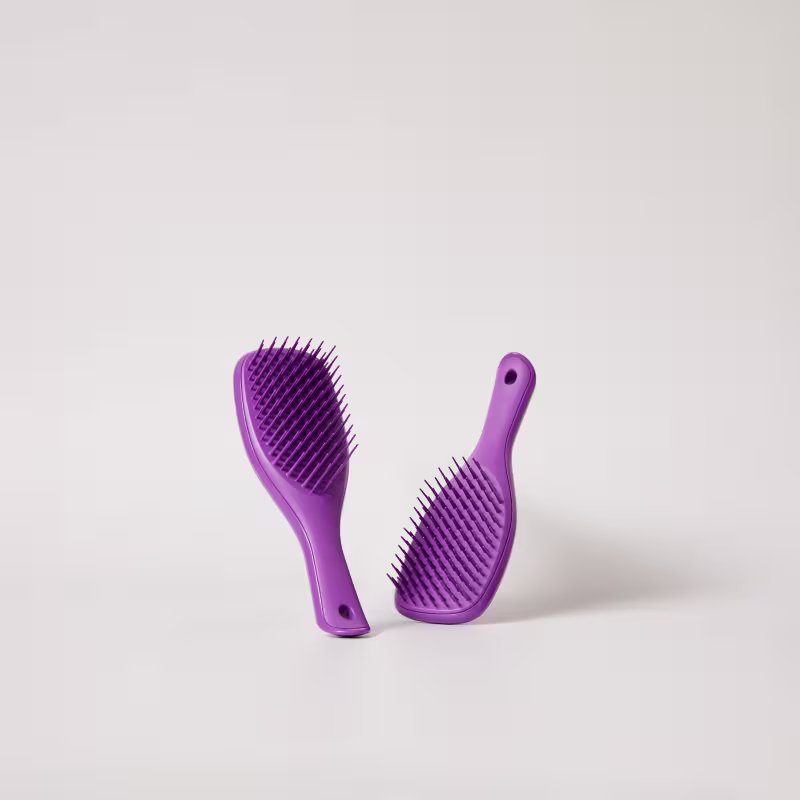 Dois pentes de cabelo de plástico de cor roxa, com dentes longos e finos, dispostos em ângulo sobre um fundo branco liso. Os pentes têm um design simples e minimalista, com um formato ergonômico e alças para facilitar o manuseio.