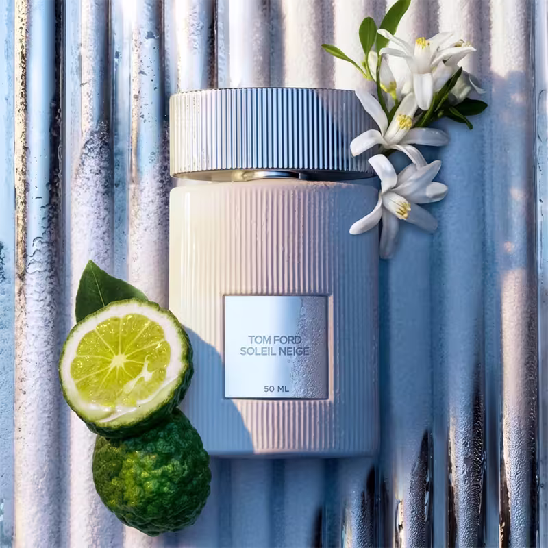 Frasco de perfume Tom Ford Soleil Neige, com embalagem branca e detalhes em azul, acompanhado de flores brancas e uma fatia de limão verde.