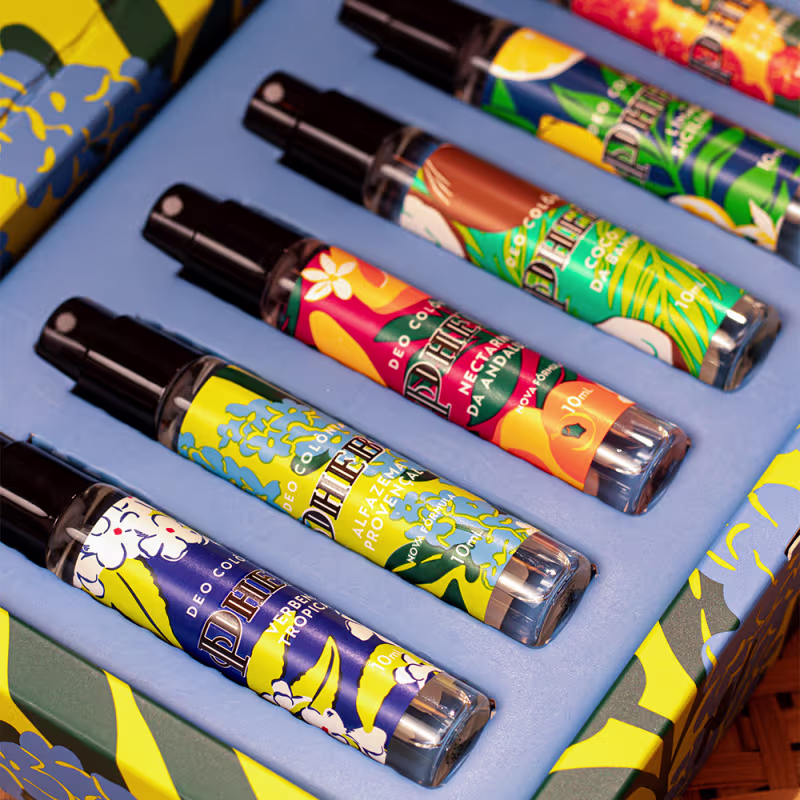Caixa amarela com flores azuis e verdes, contendo diversos frascos de itens de perfumaria da marca Phebo em diferentes cores e formatos.