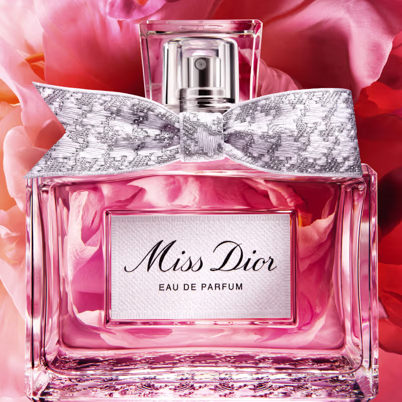 Frasco de perfume Miss Dior em formato retangular com tampa prateada. Embalagem com detalhes em renda branca e rosa claro, com texto "Miss Dior Eau de Parfum" em destaque.