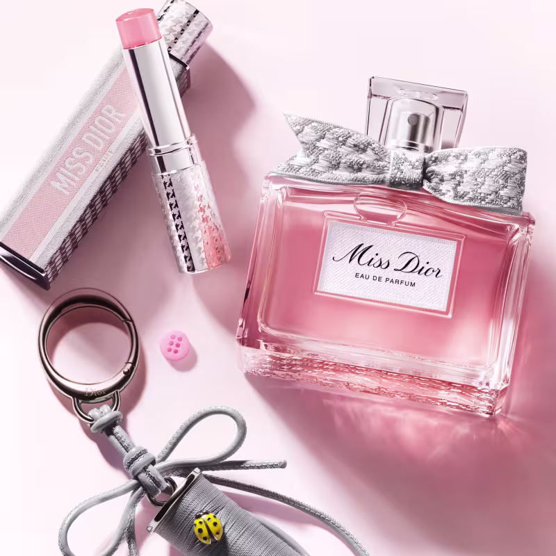 Perfume Miss Dior, batom e acessórios em tons de rosa e cinza, sugerindo um contexto elegante e sofisticado.