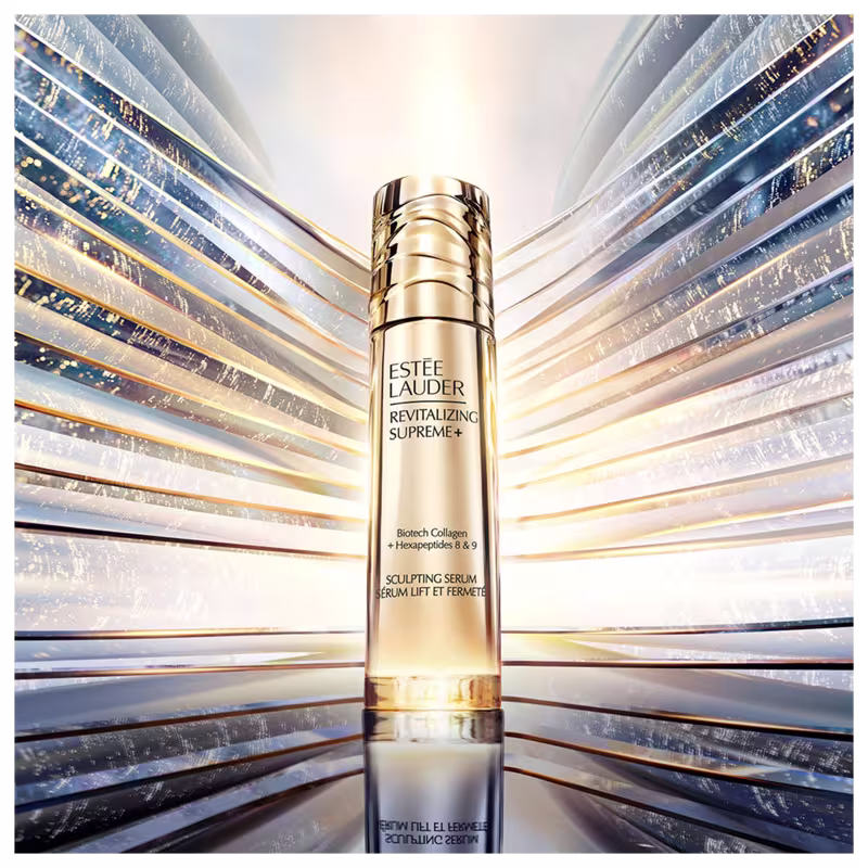 Frasco dourado da linha Revitalizing Supreme+ da Estée Lauder, contendo um soro de colágeno e hexapeptídeos para esculpir e firmar a pele. O produto é apresentado em um ambiente de luzes e reflexos brilhantes.