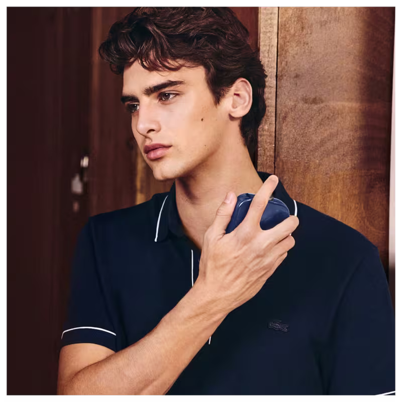 Frasco de perfume Lacoste de cor azul escuro, com textura de couro e logotipo da marca em destaque. Embalagem elegante e minimalista, com tampa preta.