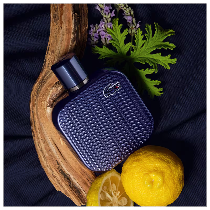 Frasco de perfume Lacoste de cor azul escuro, com textura de grade e logotipo da marca em destaque. Embalagem cilíndrica com tampa preta, apresentando design elegante e sofisticado.