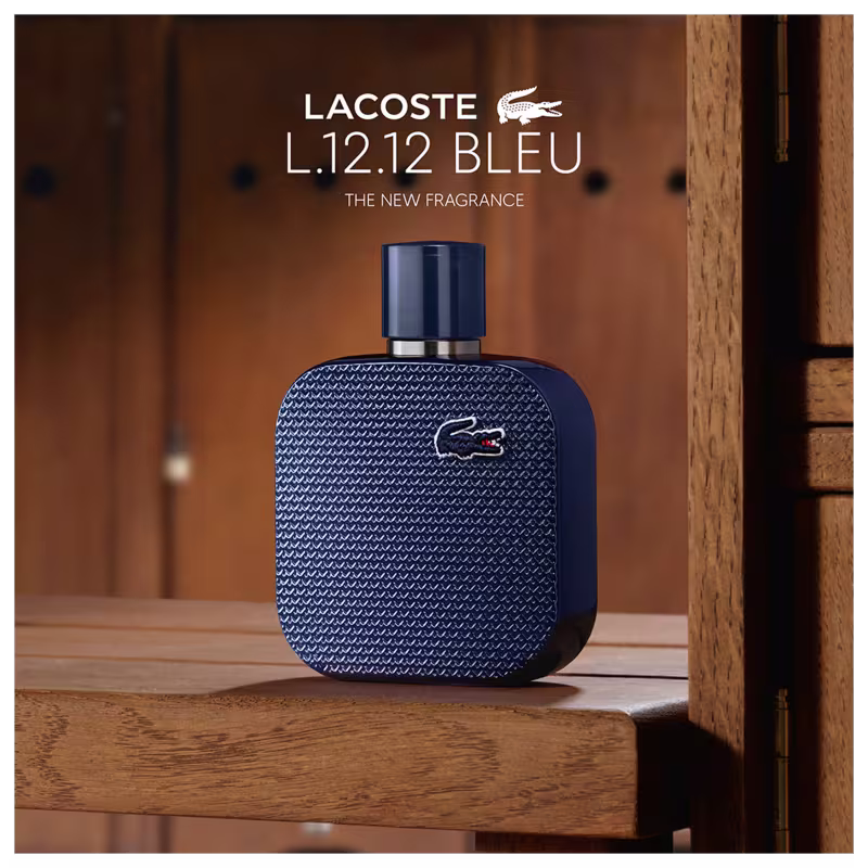 Frasco de perfume Lacoste de cor azul escuro, com textura de grade e logotipo da marca em destaque. Embalagem cilíndrica com tampa preta, apresentando design elegante e sofisticado.