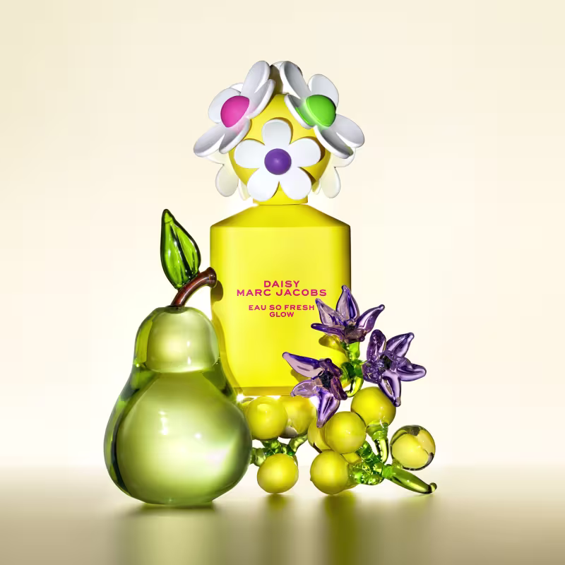 Frasco de perfume Daisy Marc Jacobs Eau So Fresh Glow em embalagem amarela com flores brancas, verdes e roxas decorativas. Pêra verde e outros elementos florais complementam a composição.