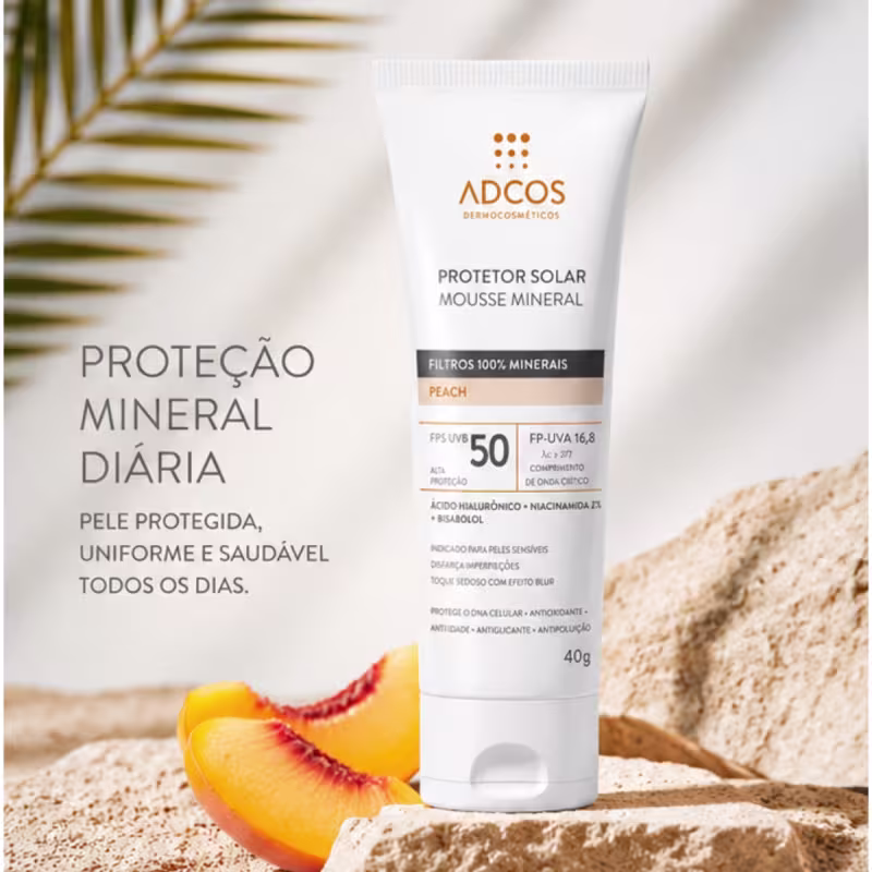 Ambiente de praia com areia, destacando um protetor solar mineral da marca Adcos. Texto: Proteção mineral diária. Pele protegida, uniforme e saudável todos os dias. Filtros 100% minerais. FPS 50. Ácido hialurônico + niacinamida. Bisabolol.