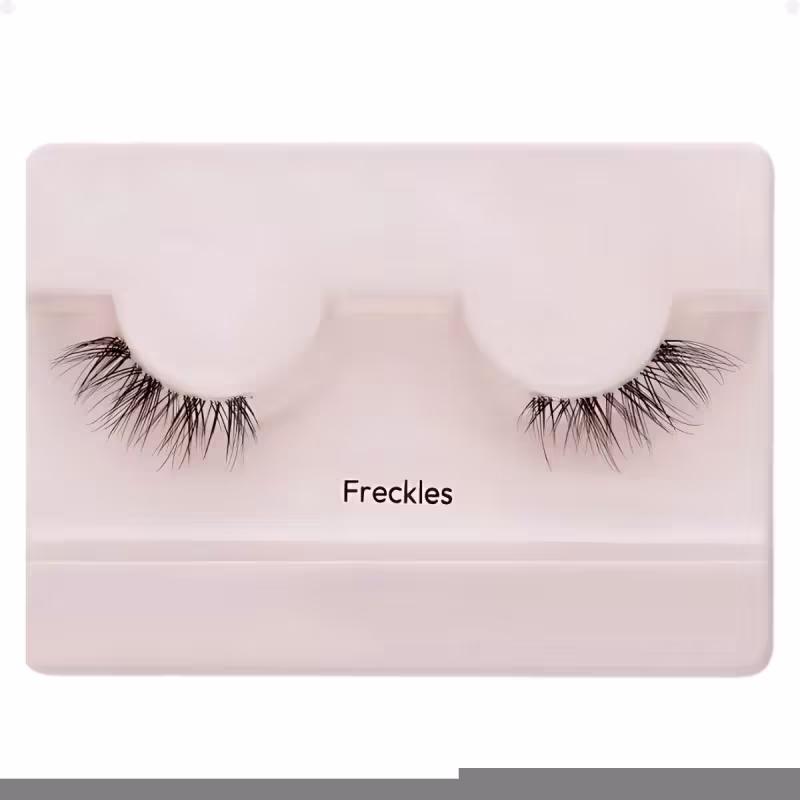 A imagem mostra os cílios postiços em um estojo plástico claro. O estojo tem uma cor nude claro e exibe um par de cílios postiços pretos.