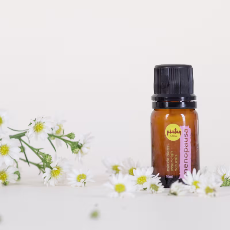 Frasco âmbar de óleo essencial com o nome "menopausa" escrito. Ao redor do frasco, há pequenas flores brancas com centros amarelos espalhadas, aparentando ser  camomilas.