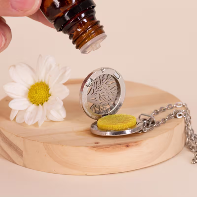 A imagem mostra o óleo sendo pingando em um pingente de metal prateado em forma de folhas com pedra amarela. Sobre base de madeira clara, acompanhado de flor branca com miolo amarelo.