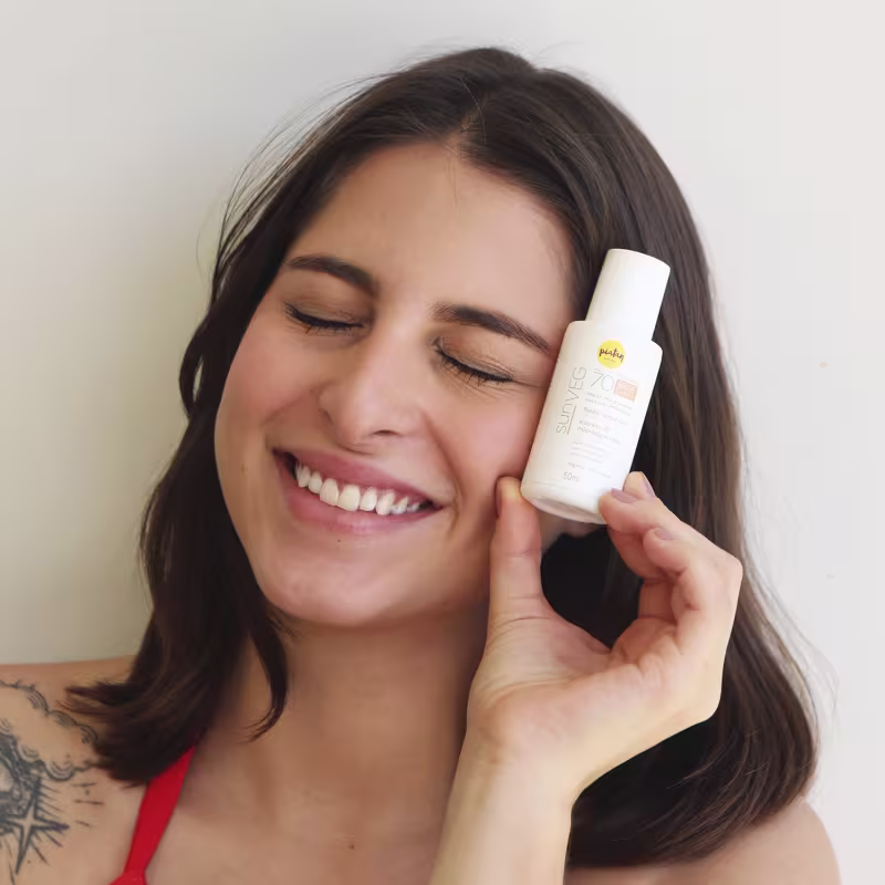 A imagem mostra uma mulher branca jovem sorrindo com os olhos fechados e segurando o protetor solar ao lado do seu rosto.