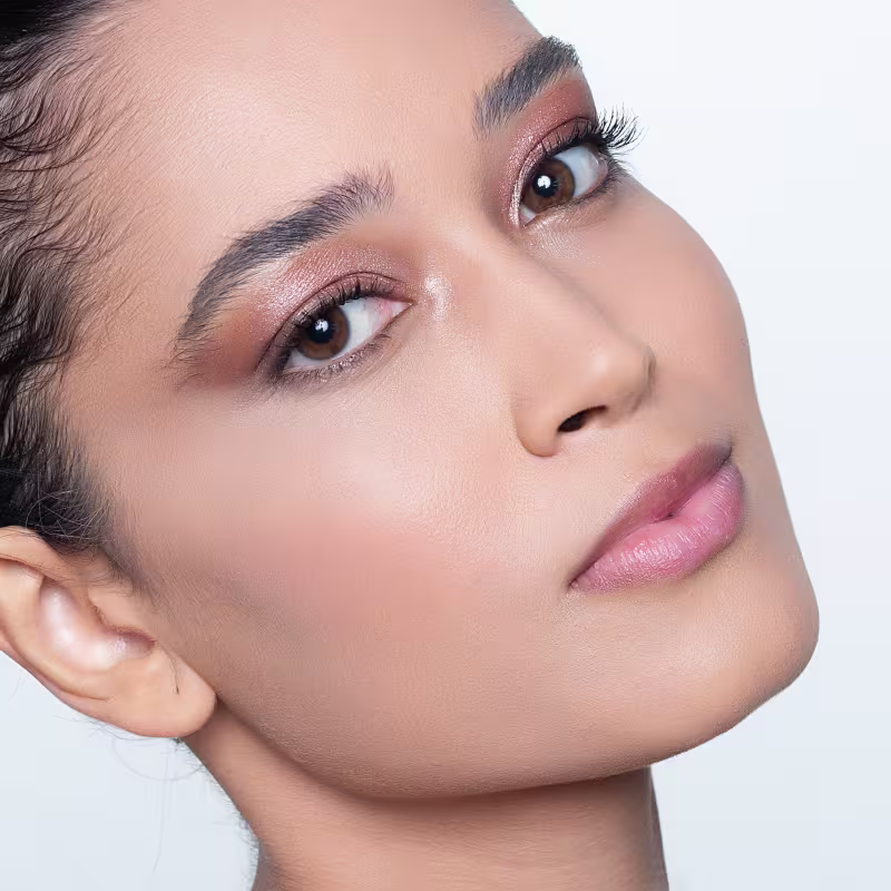 Modelo de pele clara com olhos bem delineados com maquiagem marrom e cílios longos e curvados. A pele tem um aspecto natural e iluminado. Os lábios estão com um batom rosado claro, dando um toque suave e delicado.