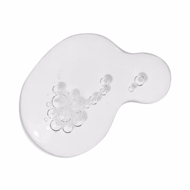 A imagem mostra algumas gotas do produto derramadas sobre uma superfície. É possível ver uma textura aquosa transparente, com pequenas bolhas de ar.