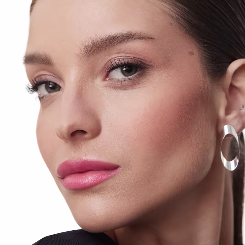 A imagem mostra um close-up do rosto de uma mulher usando maquiagem. Ela tem sobrancelhas bem delineadas em um tom castanho escuro. Seus olhos estão realçados com sombra marrom delineador e cílios volumosos. Os lábios estão com um batom rosado brilhante.