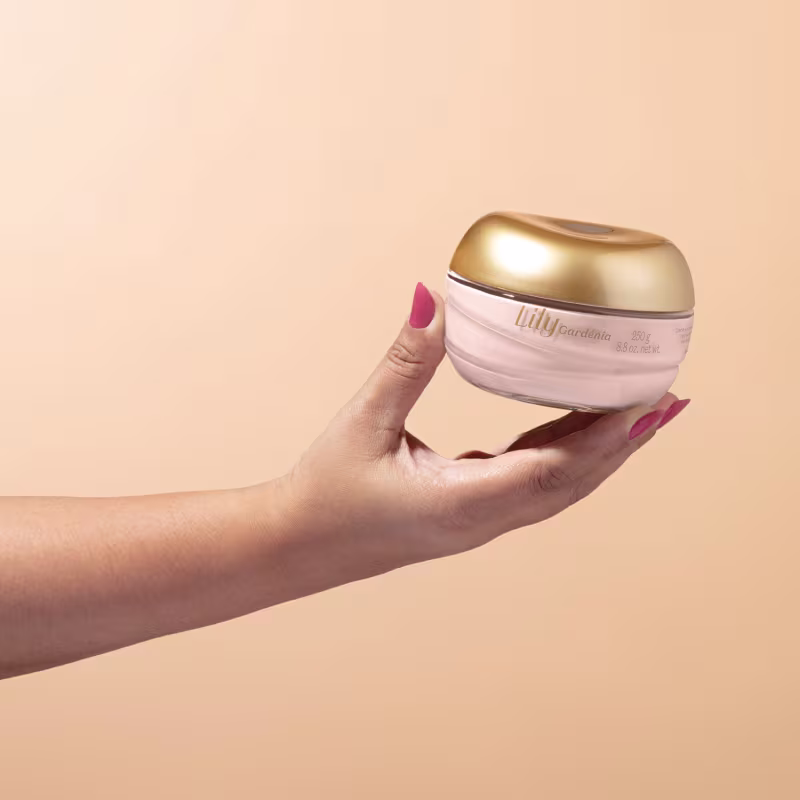 Produto de cuidados com a pele Lily Gardenia. Frasco arredondado de plástico branco com tampa dourada metálica brilhante. O frasco é segurado por uma mão feminina com unhas pintadas de rosa contra um fundo bege claro.