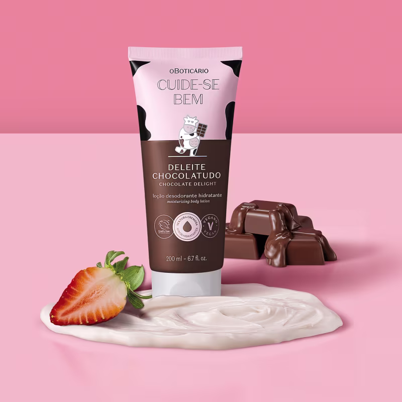 Loção hidratante corporal com aroma de chocolate. Imagem mostra a embalagem ao lado de barras de chocolate, uma fatia de morango e um creme rosa claro, todos dispostos sobre um fundo rosa. Texto em português indica que é uma loção hidratante com aroma