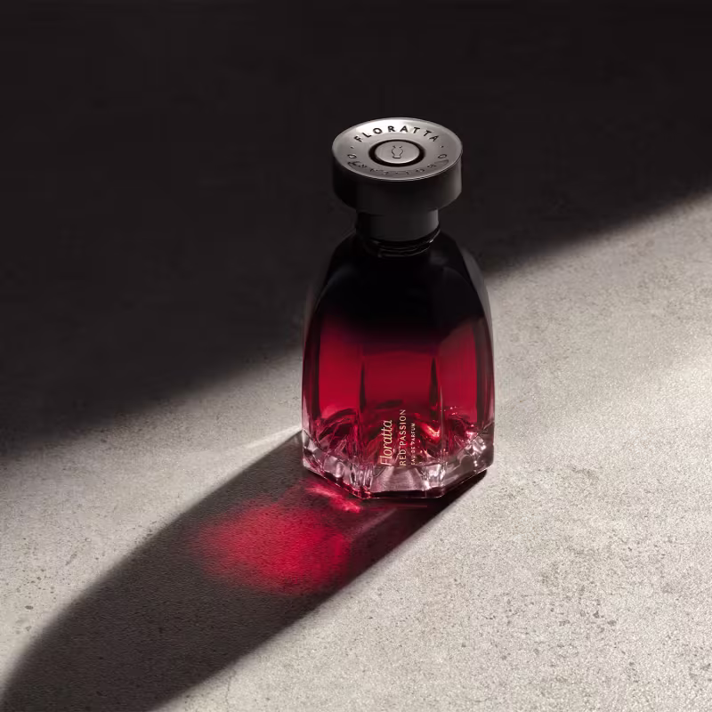 O produto apresentado é um frasco da marca Floratta Red Passion. O frasco possui um design geométrico com facetas, e a coloração do líquido varia de um tom escuro próximo ao preto até um vermelho intenso.