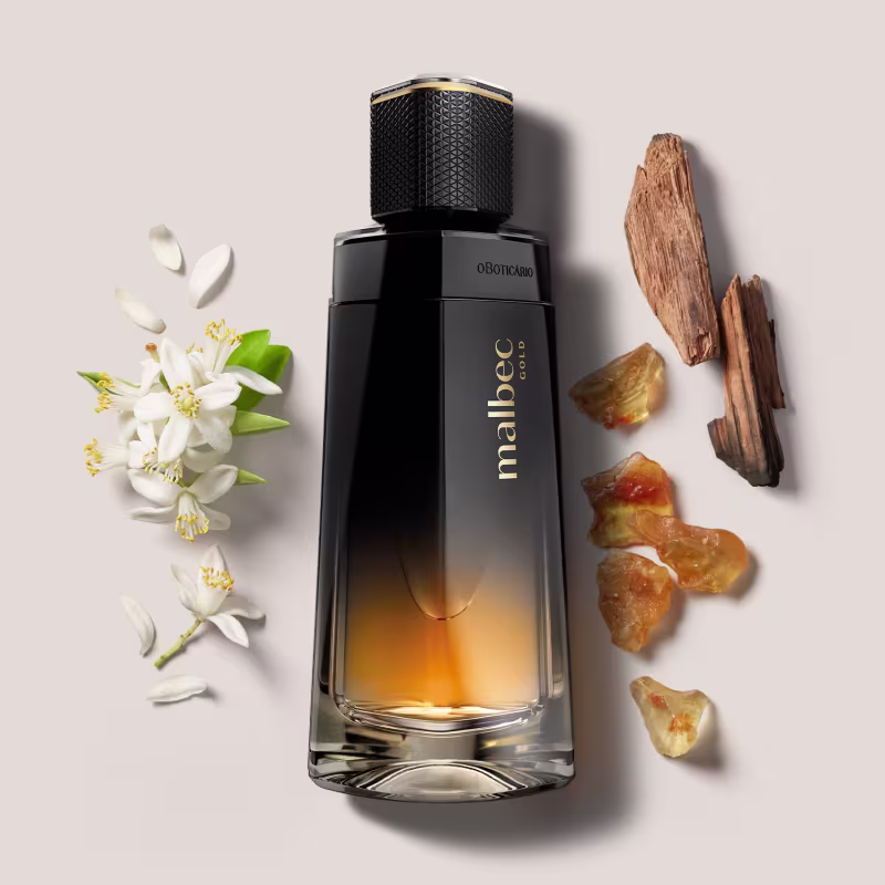 A fragrância parece ter uma coloração âmbar, sugerindo uma fragrância amadeirada ou amadeirada-floral. Ao redor do frasco, há alguns elementos naturais, como flores brancas e pedras.