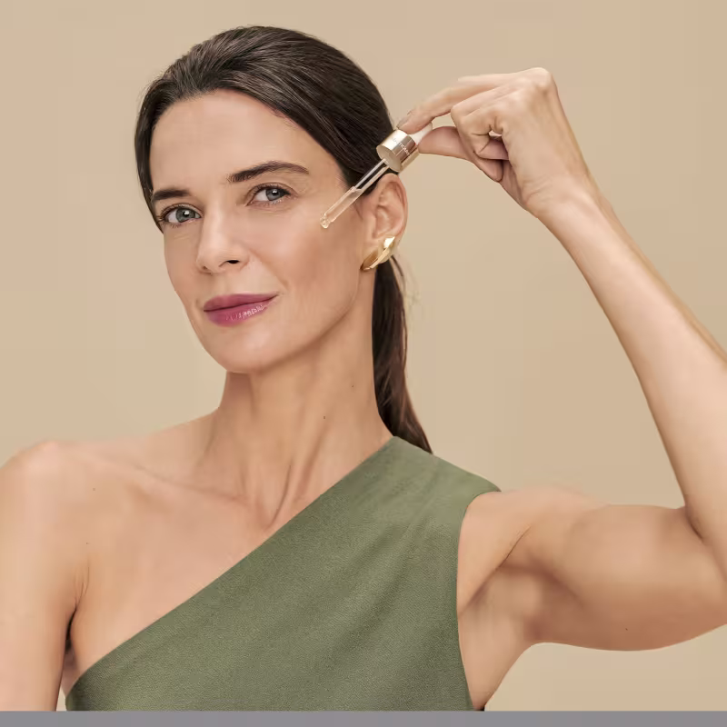A mulher aplica um produto de beleza em seu rosto em um ambiente neutro de tons claros, sugerindo um contexto de cuidados com a pele e manutenção da aparência.