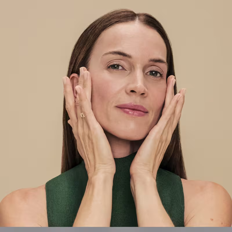 Ambiente de beleza com mulher madura em primeiro plano, usando suéter verde e expressão pensativa, em cenário bege neutro, sugerindo um contexto de cuidados com a pele e rejuvenescimento da marca Botik.