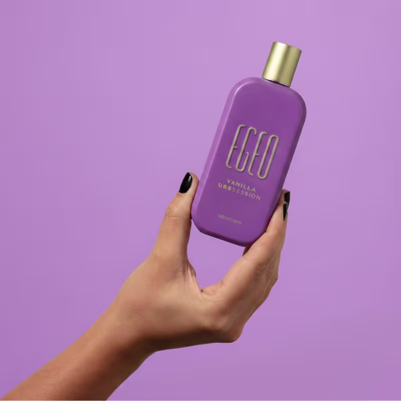 Frasco de item de perfumaria da marca Egeo em tom de roxo, com tampa dourada, segurado por mão feminina sobre fundo liso roxo.