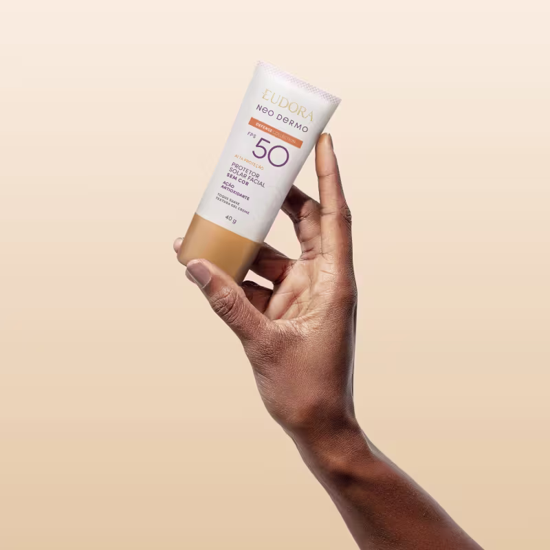 O produto mostrado está sendo segurado por uma mão, é um protetor solar Neo Dermo com FPS 50, em um tubo branco com detalhes roxo e laranja.