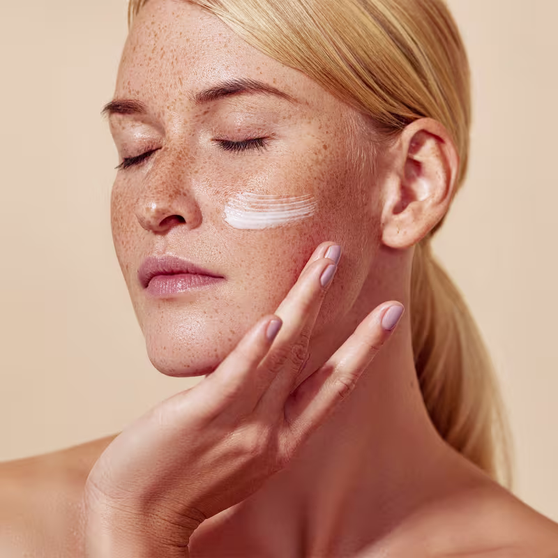 O produto (protetor solar) está sendo aplicado no rosto de uma pessoa, com uma textura branca e cremosa visível. A embalagem não é mostrada, mas o produto parece estar sendo aplicado diretamente na pele.