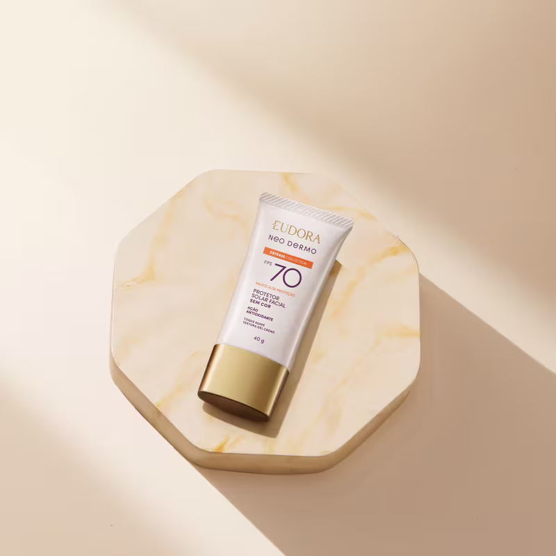 O produto apresentado na imagem é um protetor solar da linha Neo Dermo da marca Eudora. Trata-se de um tubo de cor branca e dourada, com o rótulo indicando um fator de proteção solar 50. O produto é descrito como um "Defensor Solar" que protege a pele.