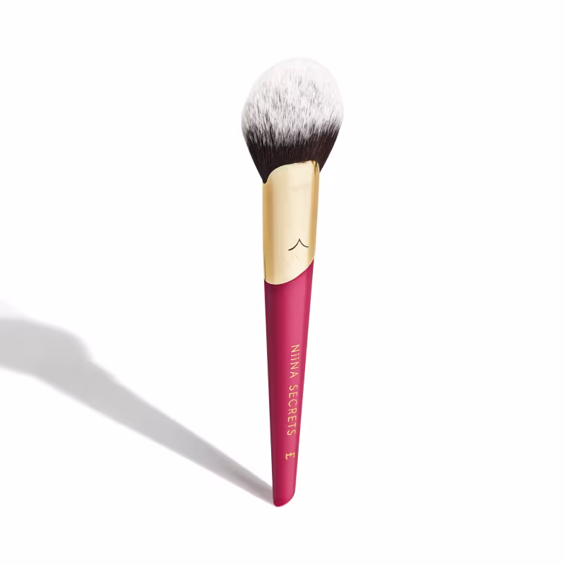 Pincel de maquiagem com cabo rosa e parte superior dourada. O cabo é cilíndrico e tem a inscrição "Niina Secrets" em dourado. As cerdas do pincel são densas, com tonalidades que vão do branco ao marrom escuro em formato arredondado uniforme.