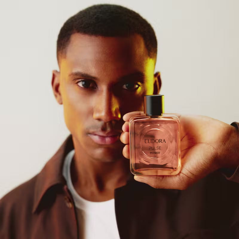 O produto mostrado na imagem é um frasco do Desodorante Colônia Pulse Power. O frasco possui um design moderno e elegante, com uma tonalidade rosada predominante. O produto é segurado por um modelo que usa uma camiseta branca e uma jaqueta marrom.