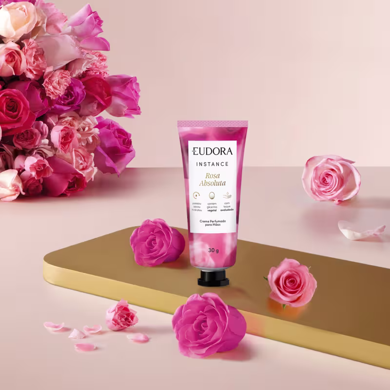 O creme está em uma embalagem de tubo plástico rosa, com o logo da marca e informações sobre os ingredientes. O produto está posicionado em uma superfície dourada, com algumas rosas frescas e pétalas espalhadas ao redor, criando uma composição floral.