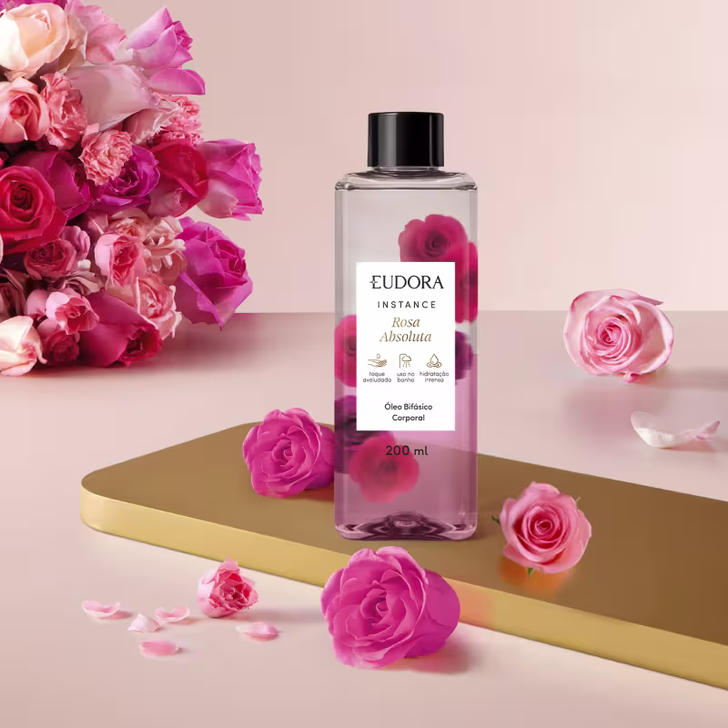 O frasco do Óleo Bifásico Instance Rosa Absoluta é transparente, com um rótulo preto e branco, e contém um líquido claro. O produto está posicionado em um ambiente com rosas rosa, criando uma composição visual harmoniosa.