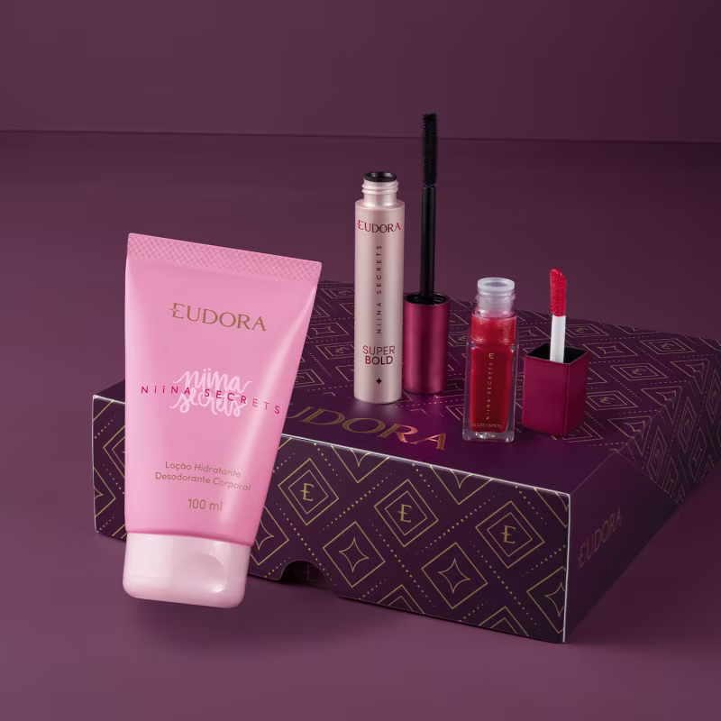 A caixa roxa e dourada da marca Eudora contém diversos produtos de beleza, incluindo uma loção hidratante corporal da linha Nina Secrets, um gloss que está aberto e um frasco de máscara de cílios preta também com o aplicador ao lado.