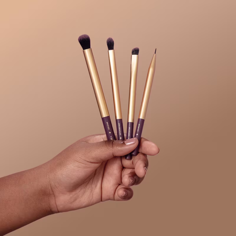 Mão segurando quatro pincéis de maquiagem da marca Eudora. Os pincéis possuem hastes douradas e cerdas escuras. Eles variam em tamanho, com diferentes formatos de ponta, indicando diferentes usos.